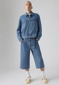 Chaqueta de denim y culottes a juego en azul claro. Chaqueta con dos bolsillos en el pecho, detalles desgastados y abertura frontal. Zapatillas y calcetines blancos.