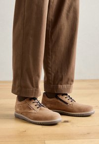 Zapatos de ante marrón con cordones, con un acabado texturizado, presentan cordones azul oscuro y suela de goma, combinados con pantalones marrones.