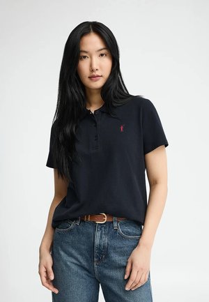 Jonge vrouw met lang zwart haar, gekleed in een zwart poloshirt en blauwe spijkerbroek met een bruine riem, staand tegen een effen witte achtergrond.