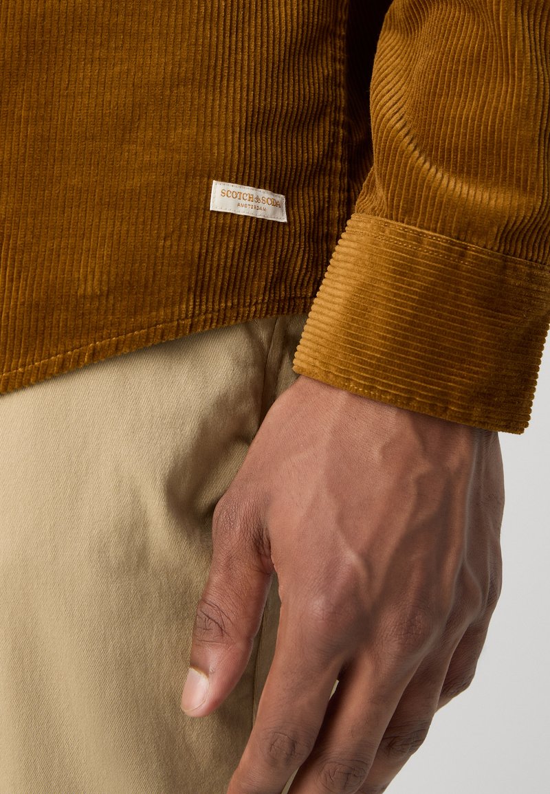 Braunes Cordhemd mit ausgeprägter Ripp-Textur, gesticktem Saum und einem Etikett mit "Scotch & Soda, Amsterdam" in der unteren Ecke.