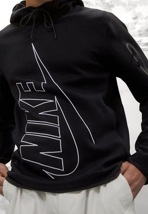 Personne portant un sweat à capuche Nike noir avec un grand logo blanc contour sur le devant et un pantalon blanc, debout, les mains dans les poches.