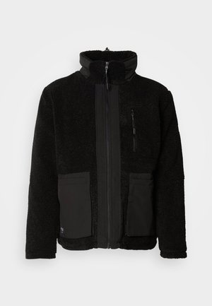 Chaqueta de forro polar negra con cuello alto, cremallera frontal y dos grandes bolsillos de parche. Presenta acentos de textil suave en la cremallera y en los bolsillos.