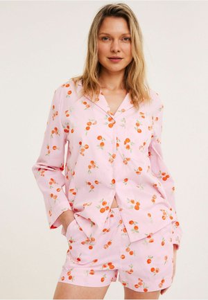 Vrouw draagt lichtroze pyjama met oranjebladerenprint, een lange mouwen blouse met knopen en bijpassende shorts, staand met handen in de zakken.