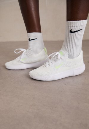 Valkoiset Nike-tennarit, joissa on teksturoitu verkkomateriaali ja neonvihreät yksityiskohdat, yhdistettyinä ribbikuvioisiin valkoisiin sukkiin, joissa on musta logo.