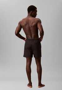 Zwarte zwembroek met een elastische tailleband en zijzak, getoond op een persoon van achteren, tegen een effen grijze achtergrond.