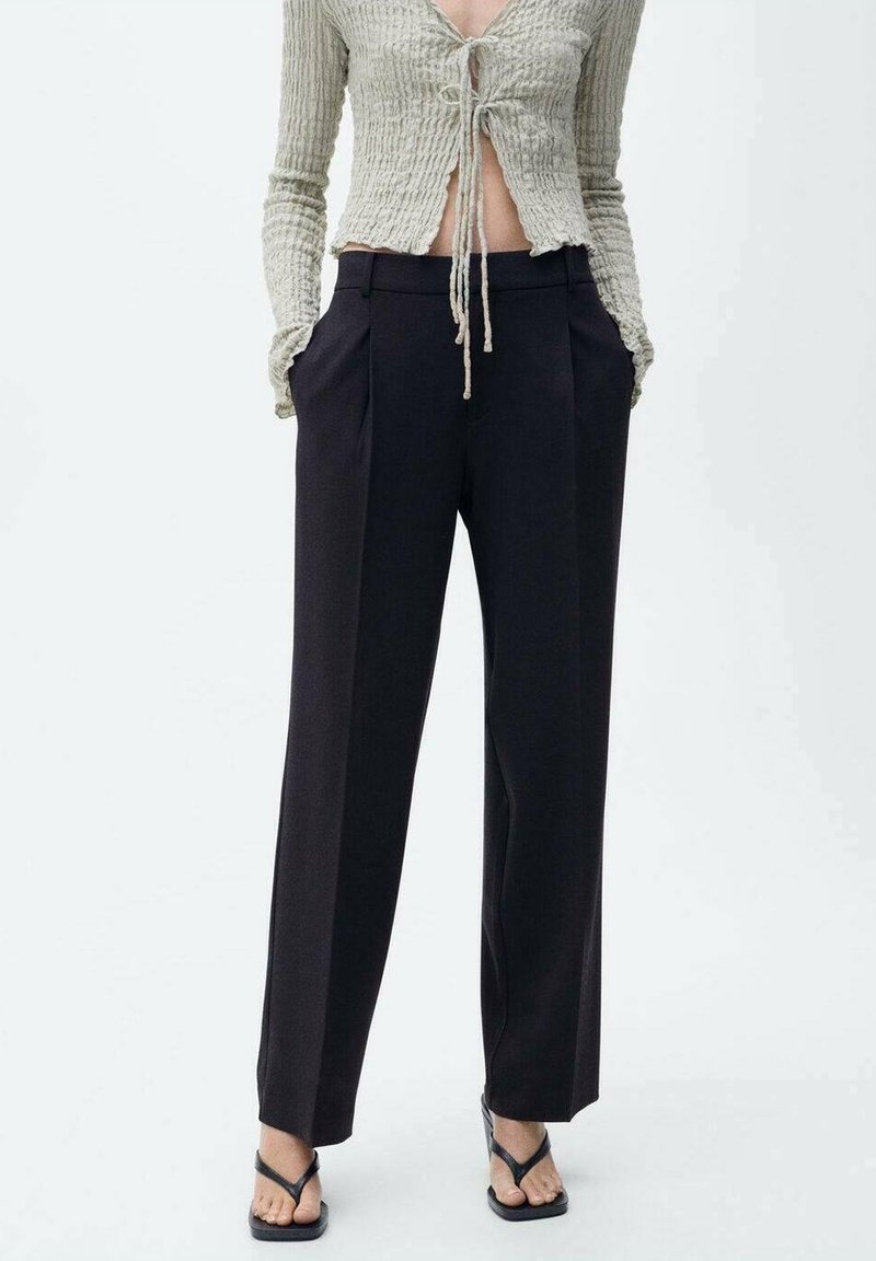 Pantalon noir slim taille haute porté avec des sandales noires à bout carré et un top court beige côtelé avec des liens à l'avant.