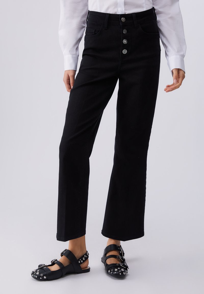 Personne portant un pantalon noir taille haute avec quatre boutons décoratifs à l'avant et des sandales noires cloutées, portant une chemise blanche à manches longues visible.