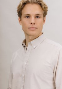 Beige skjorte med knapper laget av bomull, med klassisk krage og subtil tekstur. Modellen poserer foran en nøytral bakgrunn.