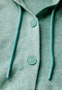 Mintgrön hoodie med två stora knappar, en slät dragkedja och matchande snoddar. Tyget är mjukt, strukturerat och något melerat.