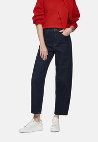 Dunkelblaue Jeans mit hoher Taille und geradem Bein, kontrastierende Nähte und Seitentaschen, kombiniert mit weißen Sneakern. Roter Pullover mit strukturierter Oberfläche.