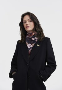 Juodas paltas su pritaikytu dizainu, derinamas su tamsių tonų gėlių raštuotis šaliku. Šaliką puošia mažas dekoratyvinis segtukas.