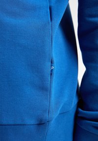 Sweat-shirt bleu en tissu doux et texturé, avec des poignets côtelés et une petite étiquette logo sur la couture latérale. Coupe standard avec une finition lisse.