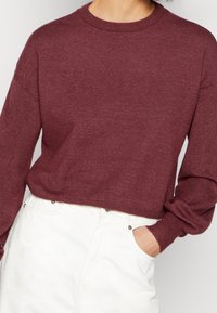 Osoba ubrana w bordowy, długi sweter dzianinowy włożony w białe spodnie w wysokim stanie, z jedną ręką w kieszeni.