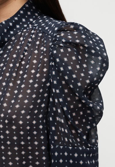 Κοντινό πλάνο ενός διαφανούς μπλε μπλουζιού με φουσκωτά μανίκια και λευκό και ροδακινί διαμαντένιο σχέδιο.