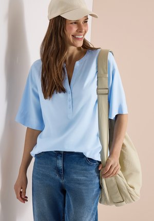 Femme portant une casquette beige, un chemisier bleu clair à manches courtes, un jean bleu, souriante et portant un sac à épaule beige.