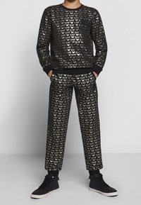 Persona che indossa una felpa nera e pantaloni con ripetuti loghi dorati "EA7", mani nelle tasche e sneakers nere alte.