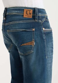 Vaqueros de mezclilla azul oscuro con un lavado medio. Las características incluyen un parche de cuero en la cintura, bolsillos laterales y costuras en contraste.