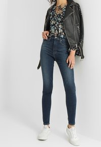 Chaqueta de cuero negra con tachuelas, blusa con estampado floral y cuello con lazo, jeans ajustados oscuros de tiro alto, y zapatillas blancas.