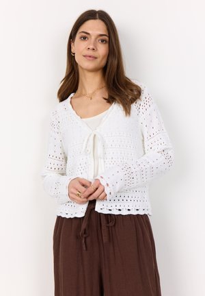 White knitted cardigan with openwork pattern, tie front closure, long sleeves, and scalloped hem. Worn over a cream top and brown pants.