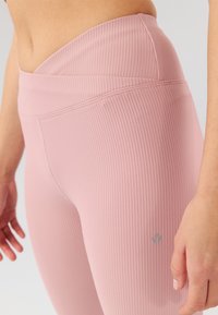 Leggings rosa chiaro a coste con vita alta, caratterizzati da un piccolo logo sul lato sinistro e una texture liscia. Dettaglio cucitura visibile.