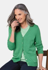 Grüne Strickjacke aus Strickstoff mit V-Ausschnitt, Knopfleiste, zwei Taschen und gerippten Bündchen. Über einem hellgrünen Oberteil getragen.
