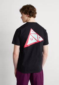 Camiseta de algodón negra con un gráfico triangular colorido en la parte posterior que presenta formas en azul claro y rosa, y el texto "HUF" dentro de los triángulos.