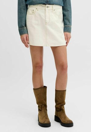 Femme portant une mini-jupe en denim blanche, une veste en denim bleue et des bottes marron en daim mi-mollet avec des talons blocs.