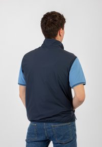 Gilet leggero blu navy con colletto alto, chiusura con zip e tasche laterali, indossato sopra una polo azzurro chiaro e jeans in denim.