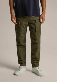 Zielone oliwkowe spodnie cargo z dwoma bocznymi kieszeniami, zwężanym krojem nogawki i materiałem bawełnianym. Noszone z białymi trampkami i granatową koszulą.