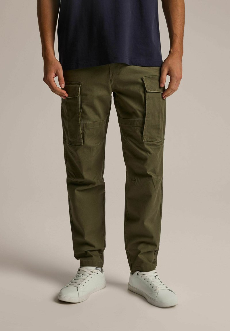 Zielone oliwkowe spodnie cargo z dwoma bocznymi kieszeniami, zwężanym krojem nogawki i materiałem bawełnianym. Noszone z białymi trampkami i granatową koszulą.