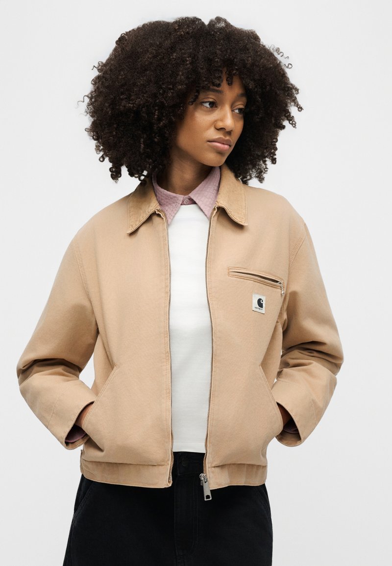 Woman with curly hair wearing a beige zip-up jacket over a white top and pink collared shirt, hands in pockets, looking to the side.