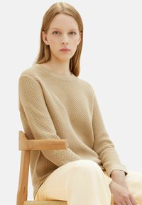 Maglione beige intrecciato con un motivo testurizzato, scollo rotondo e maniche lunghe, abbinato a pantaloni chiari, seduto su una sedia di legno.