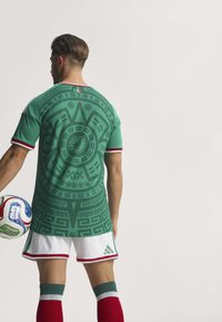 Green soccer jersey with a textured circular pattern, short sleeves with red and white accents, paired with white shorts and red socks.