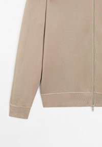 Détail des manches et de l'ourlet d'un pull en maille beige zippé avec une fermeture éclair argentée sur un fond blanc.