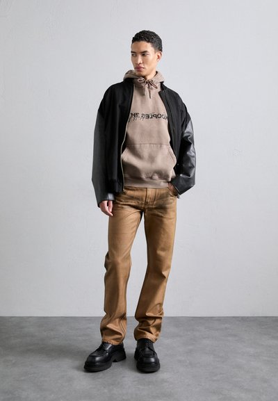 Schwarze Bomberjacke mit Lederärmel, beigefarbener Kapuzenpulli mit Logo, hellbraune Hose und schwarze klobige Schuhe. Glatte Textur, lässige Passform.