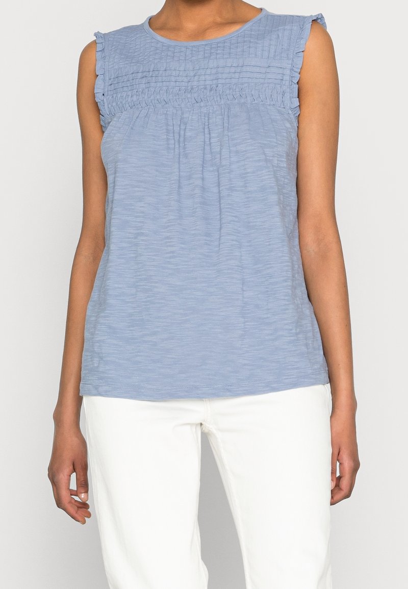 Femme portant un haut sans manches bleu clair texturé avec des épaules volantées et un pantalon blanc, debout devant un fond uni.