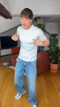 Jonge man binnen wijst naar een wit Replay T-shirt, draagt blauwe jeans en sneakers, staat op houten vloer nabij een bank en kamerplanten.
