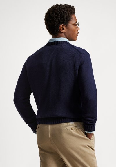Tmavě modrý pletený svetr s žebrovaným límcem a lemem, nošený přes světle modrou košili s límečkem, zobrazený zezadu.