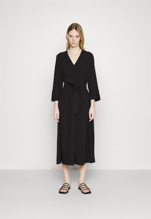 Black mid-length dress with V-neck, tied waist, and three-quarter sleeves. Smooth fabric with a flowy design, paired with black strappy sandals.