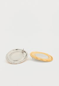 Hõbedased ja kuldsed ovaalsed taldrikud tekstuursete servadega, üks läikiv ja peegeldav, teine mati viimistlusega, mõlemal on keskel lohud.