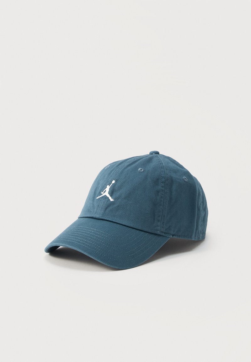 Casquette de baseball bleue avec une visière courbée et le logo Jumpman blanc à l'avant, posée sur un fond gris clair.