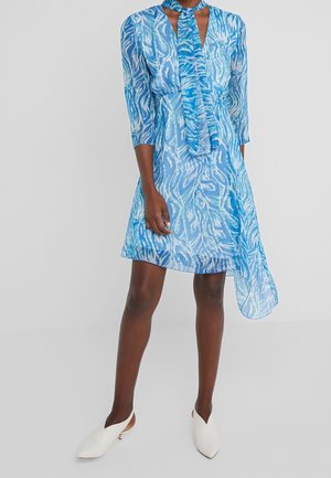 Frau trägt ein blau gemustertes knielanges Kleid mit Dreiviertelärmeln, einer Halsbinde und asymmetrischem Saum, kombiniert mit weißen spitzen flachen Absätzen.