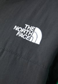 Juodas audinys su lygiu paviršiumi, su išsiuvinėtu baltu "THE NORTH FACE" logotipu ir stilizuotu ketvirtiniu apskritimu.