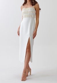 Bílé midi šaty s korálkovým třásňovým živůtkem a bočním rozparkem. Mají tenké ramínka a hladkou texturu, doplněné o nahé lodičky.