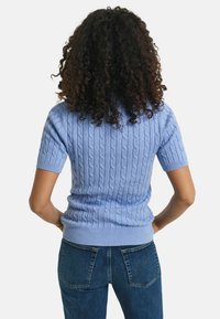 Pull court bleu clair à manches courtes avec un motif en mailles torsadées et un ourlet côtelé. Porté avec un jean bleu foncé, mettant en valeur une silhouette ajustée.