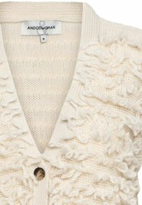 Crème-kleurige gebreide cardigan met textuur van lussen, diepe V-hals, enkele bruine knoop en merklabel "ANDCOWOMAN" maat medium.