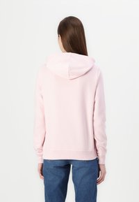 Žena s dlouhými hnědými vlasy, oblečená v svetru s kapucí světle růžové barvy a modrých džínách, viděná zezadu na jednoduchém bílém pozadí.