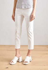 Person wearing white cropped pants and white open-toe block heel mules, standing on light wooden floor against plain background.