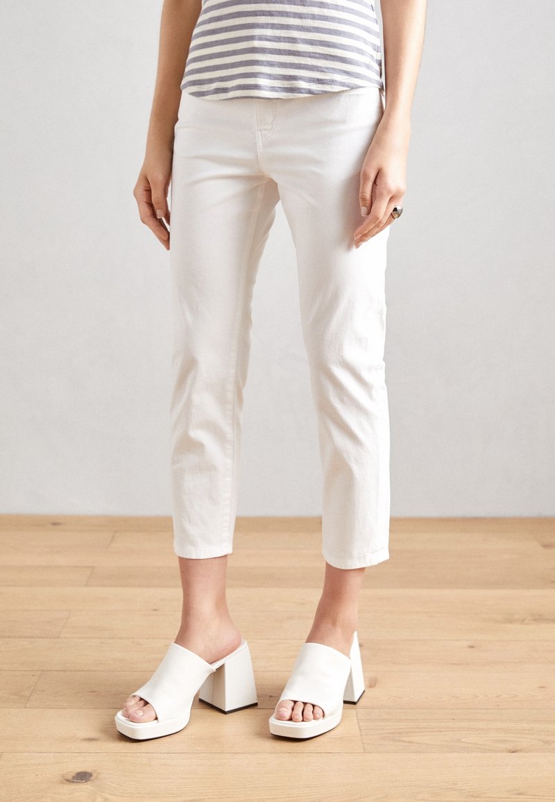 Person wearing white cropped pants and white open-toe block heel mules, standing on light wooden floor against plain background.