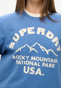 Sudadera azul con un estampado gráfico de montañas y el texto "PARQUE NACIONAL DE LAS ROCKY MOUNTAINS SUPERDRY USA" en blanco. Material de algodón.